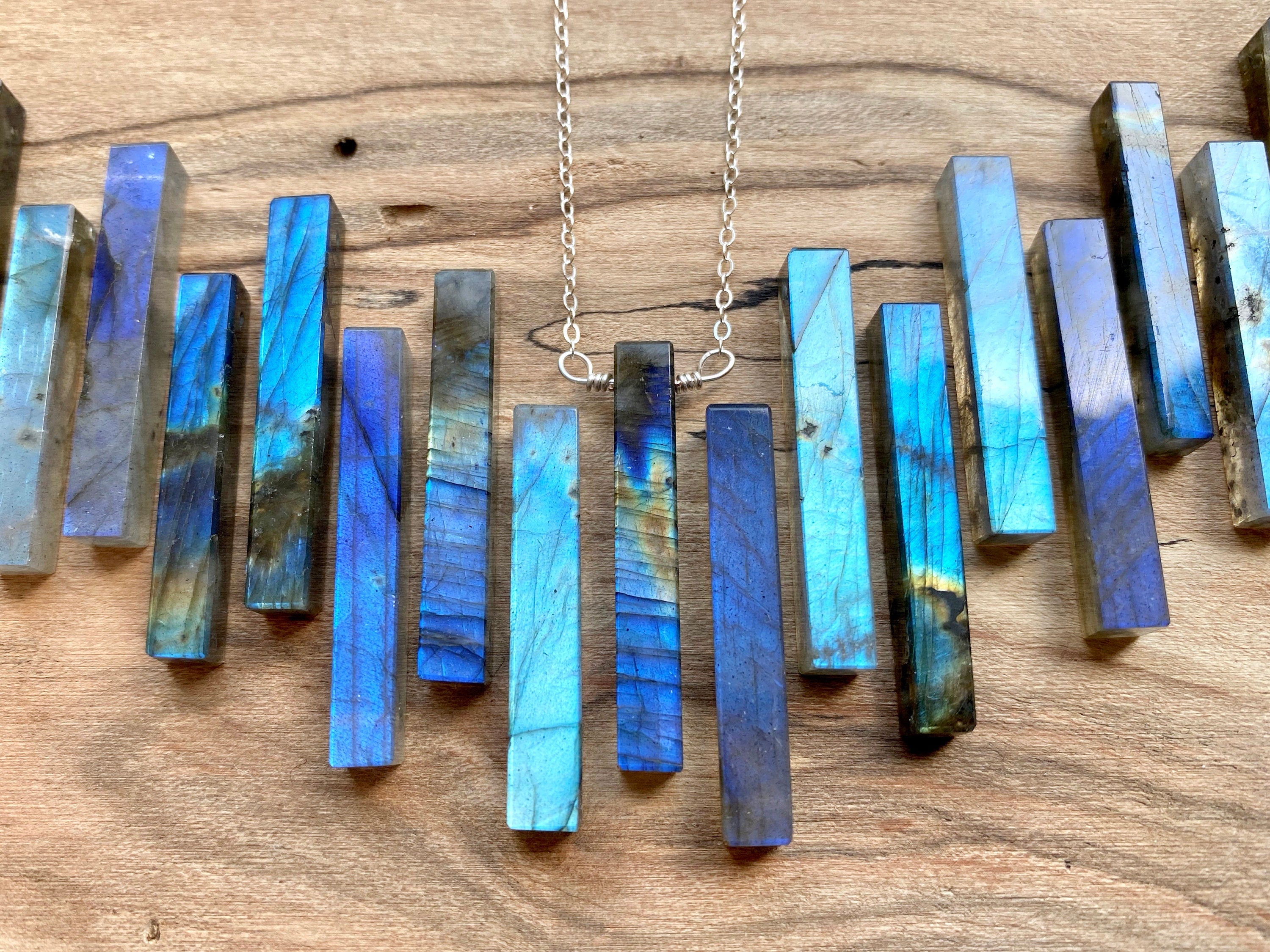 May include: A silver chain necklace with a pendant made of multiple blue labradorite stones arranged in a vertical row. The stones are rectangular and have a iridescent sheen.