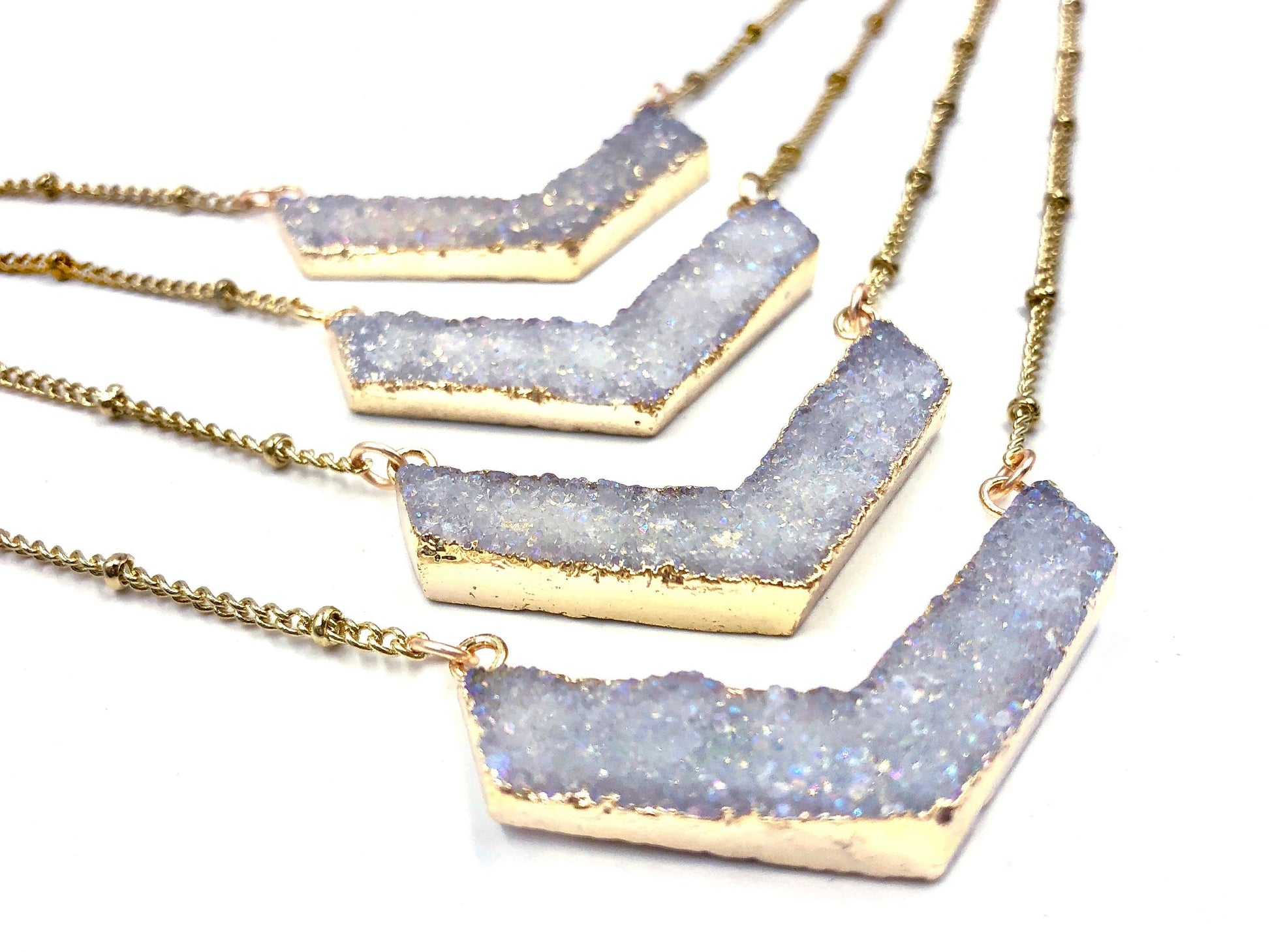 May include: Four gold chain necklaces with white crystal chevron pendants. The pendants are layered in a staggered pattern, with the largest pendant at the bottom and the smallest pendant at the top.