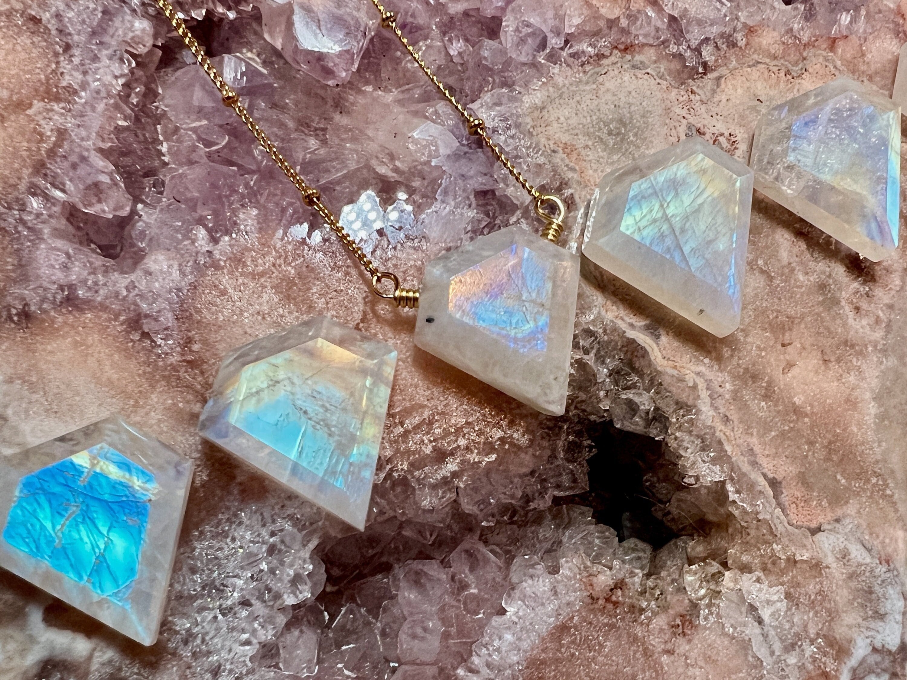 May include: Five iridescent white gemstone pendants hanging from gold chains. The gemstones are faceted and have a diamond shape. The gemstones are arranged in a row, with the top two pendants hanging from the same chain.