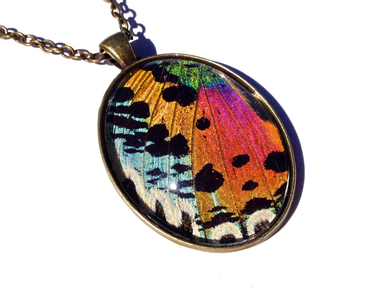 May include: A bronze oval pendant necklace with a colorful butterfly wing inside. The wing is iridescent and has shades of blue, green, orange, pink, and black. The pendant is on a chain.