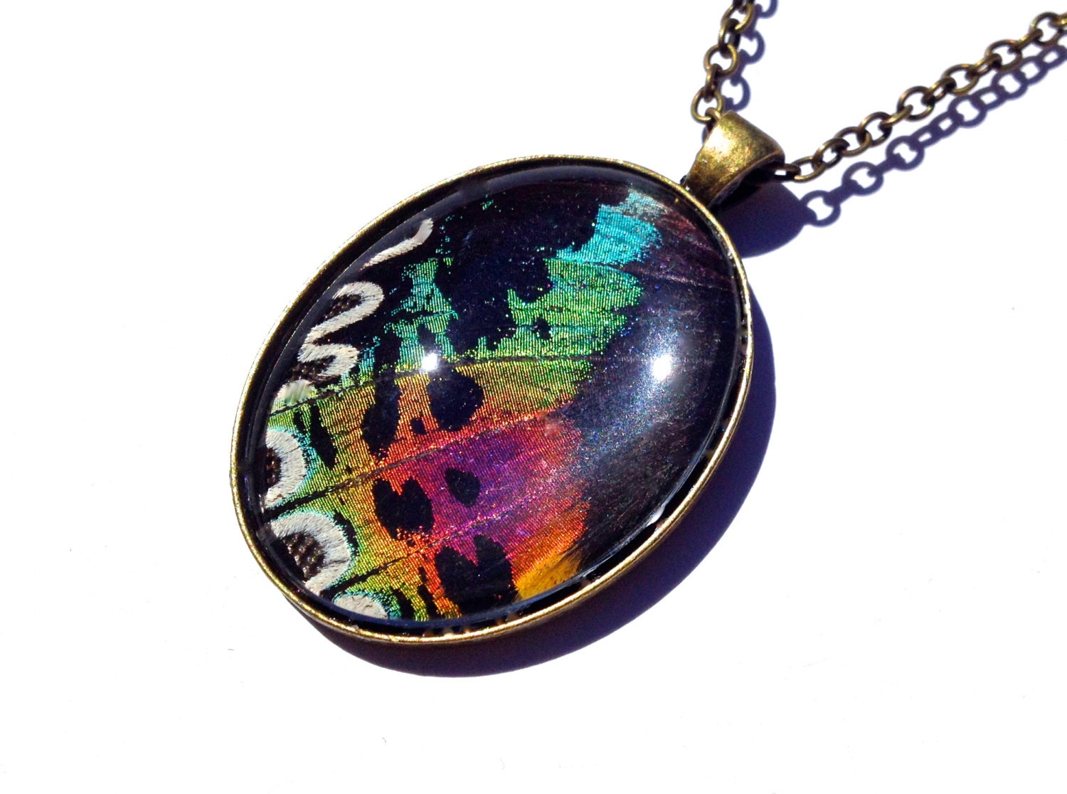 May include: A bronze oval pendant necklace with a colorful butterfly wing encased in resin. The wing has shades of green, blue, purple, pink, orange, and black.